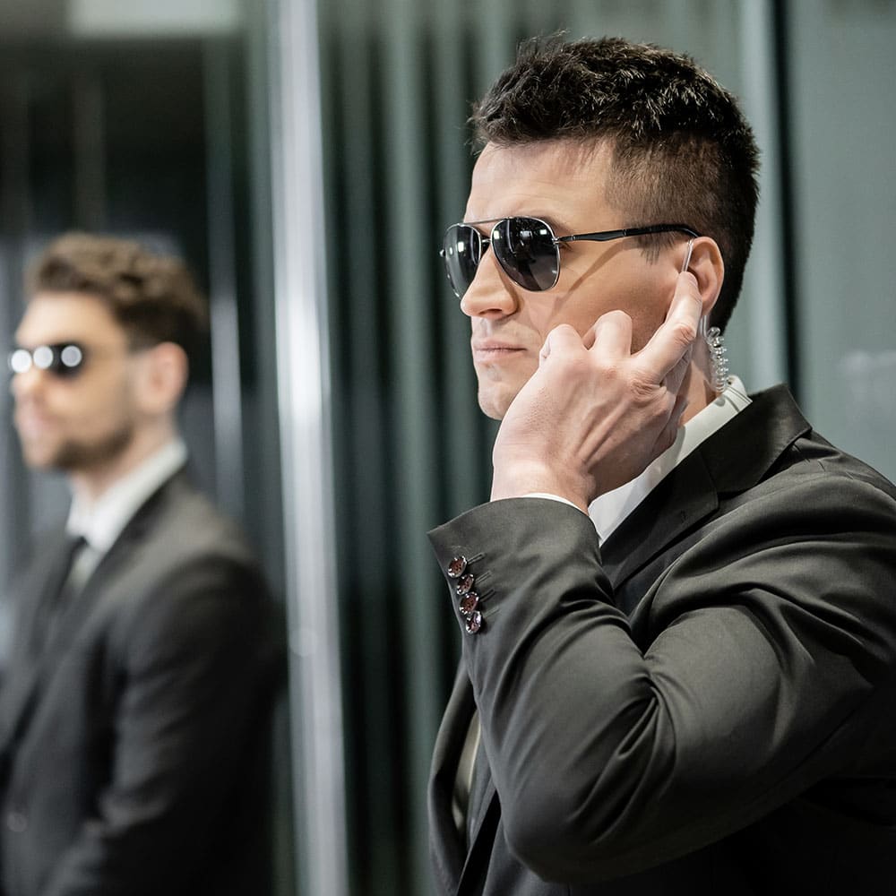 An executive protection agent wearing sunglasses adjusts his earpiece while another agent stands watch in the background, demonstrating skills learned in executive protection training.